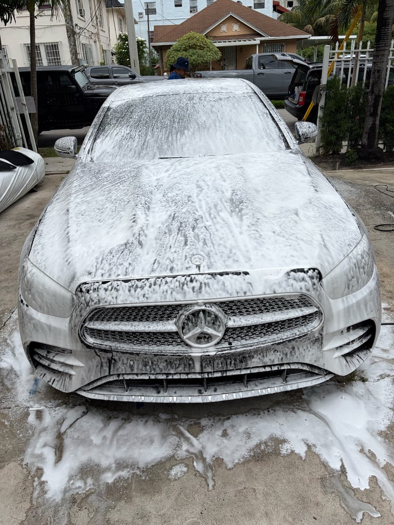 White Mercedes-Benz sedan covered in soap foam during a car wash in a residential driveway